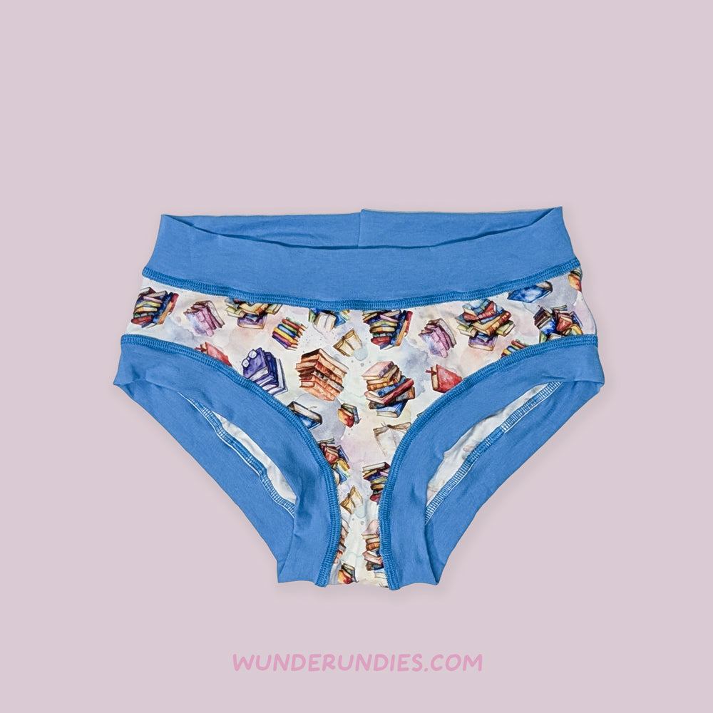 Pair of latex free underwear with colorful pattern featuring piles of books and blue waistband on a light pink background, branded 'Wunderundies'.